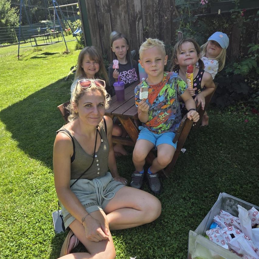 Eine Familie posiert für ein Foto im Garten. Eine Frau sitzt auf dem Rasen mit ihren Kindern um einen Tisch herum. Sie halten Eispops in den Händen.