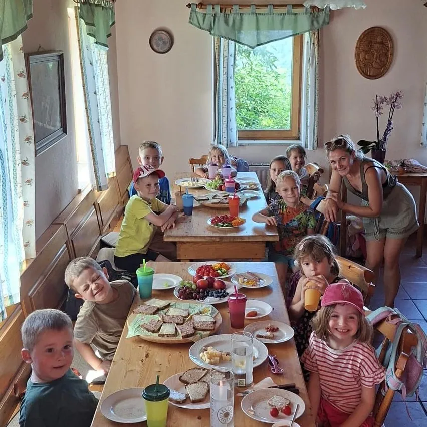 Eine Gruppe von Kindern und Erwachsenen sitzt an einem Tisch mit verschiedenen Speisen, einschließlich Obst und Sandwiches. Eine Frau steht lächelnd hinter ihnen. Der Raum hat Holzbänke und Vorhänge.