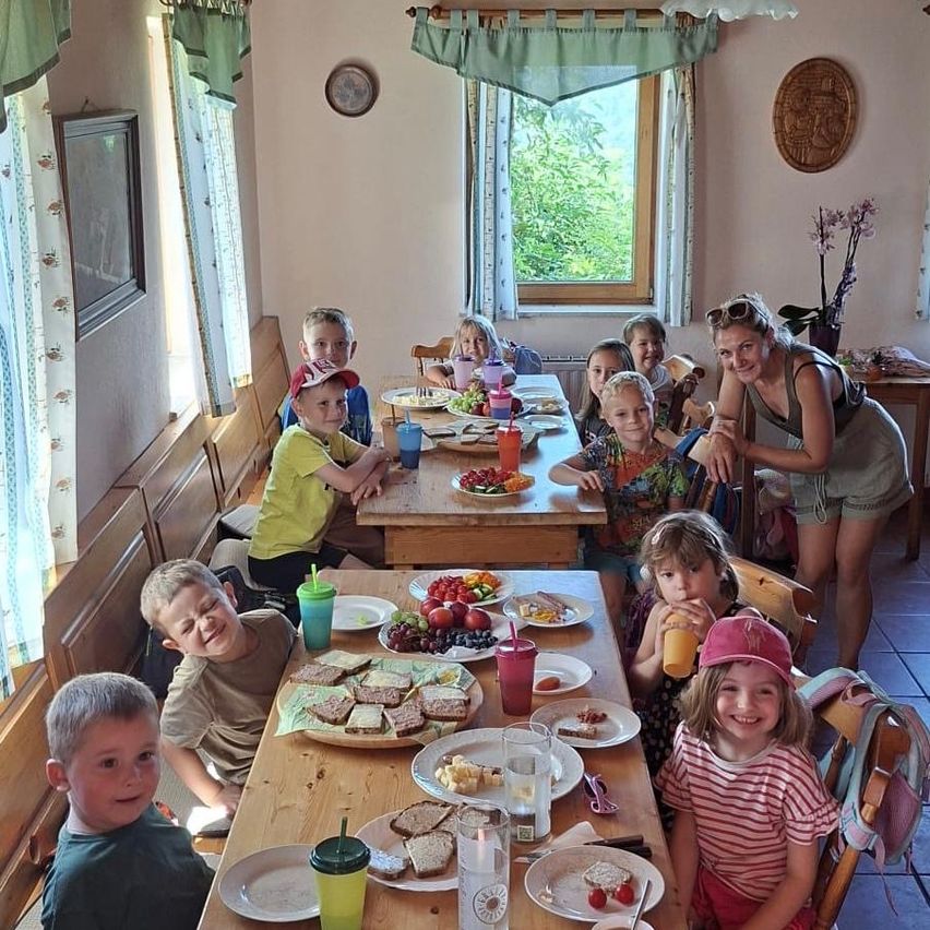 Eine Gruppe von Kindern und Erwachsenen sitzt an einem Tisch mit verschiedenen Speisen, einschließlich Obst und Sandwiches. Eine Frau steht lächelnd hinter ihnen. Der Raum hat Holzbänke und Vorhänge.