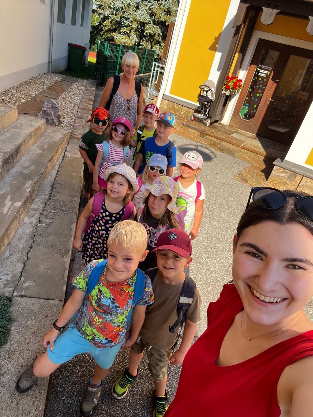 Eine Gruppe von Kindern und Erwachsenen steht vor einem Gebäude, eine Frau vorne nimmt ein Selfie auf.