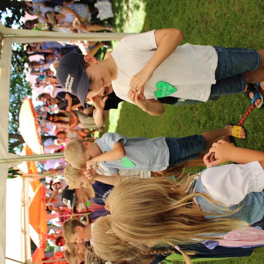 Kinder spielen im Freien auf einem Rasen. Ein Junge in einem Feuerwehrmann-Cap hält ein grünes Herz. Mehrere Kinder hinter ihm schauen etwas an. Ein Erwachsener hält ein Kind auf der rechten Seite.