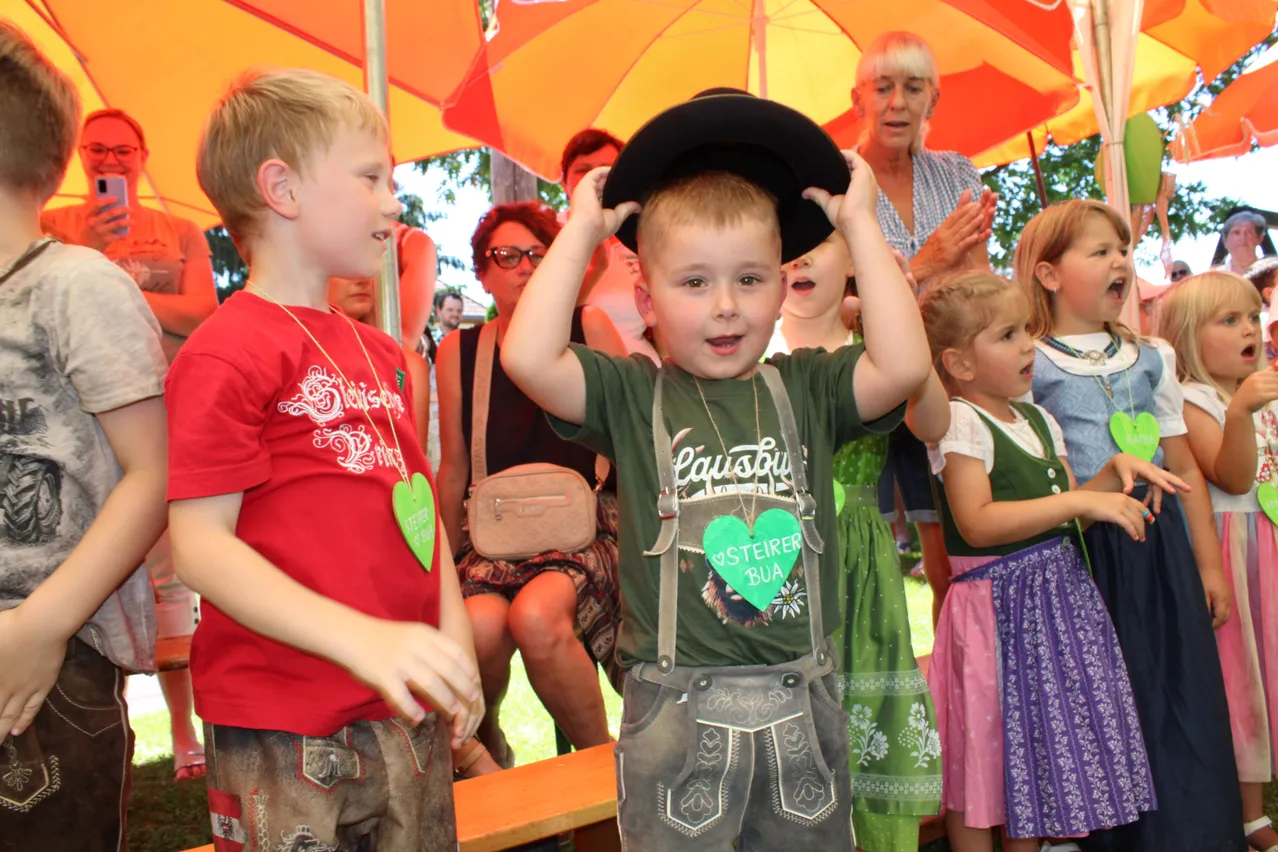 Zwei Jungen stehen unter einem bunten Schirm. Ein Junge im grünen Shirt mit Herz und Hosenträger hält seinen Hut. Eine Frau und Kinder beobachten.