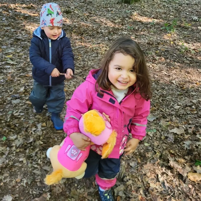 Zwei Kinder, ein Junge und ein Mädchen, gehen in einem Wald mit gefallenen Blättern. Das Mädchen hält eine Kuscheltierpuppe, während der Junge einen Stock hält.