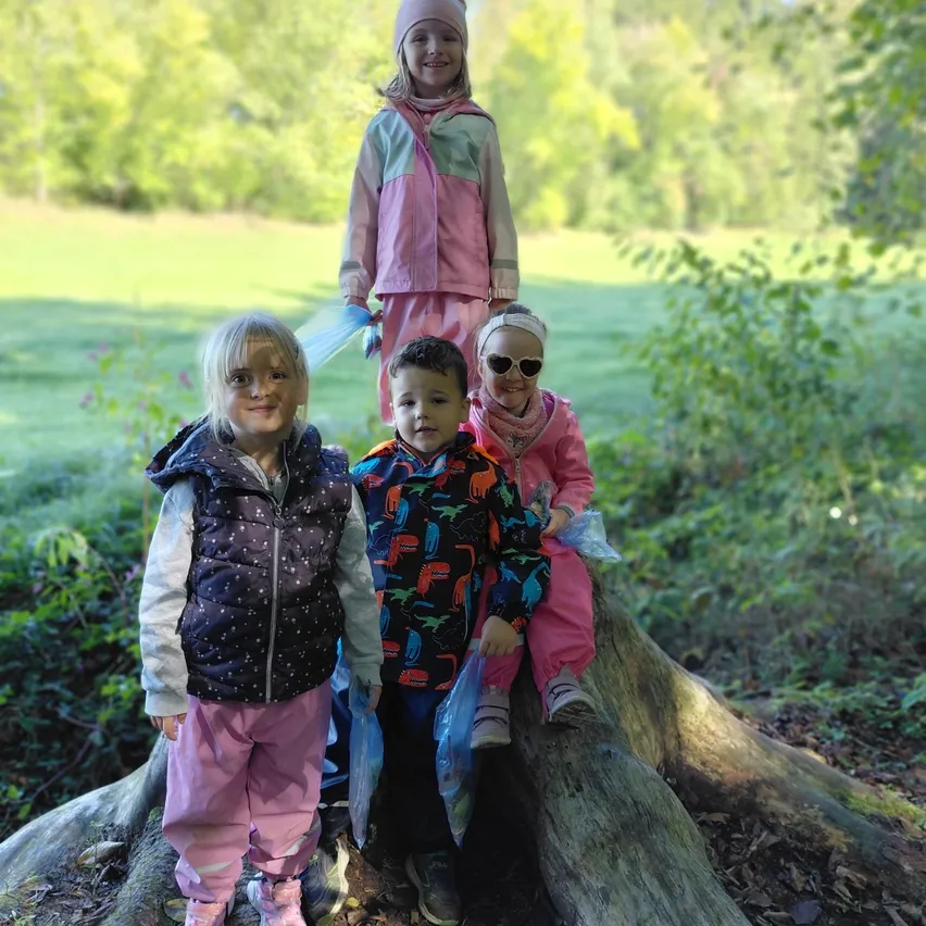 Vier Kinder stehen auf einem Baumstumpf in einem Park, lächeln und halten Plastiktüten. Ein Kind steht auf dem Stumpf, während andere auf dem Boden stehen.