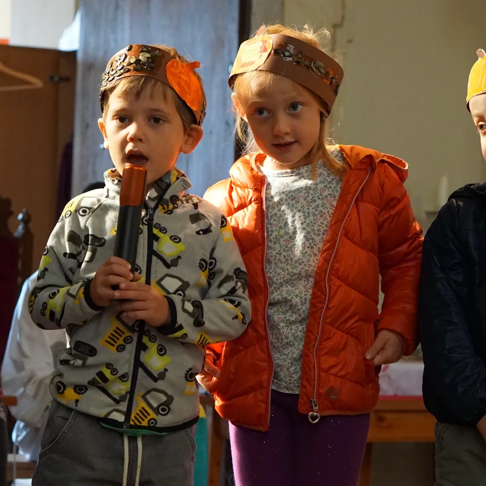 Drei Kinder stehen in einem Raum. Der Junge auf der linken Seite hält ein Mikrofon und scheint zu singen. Das Mädchen neben ihm trägt eine Kopfbinde und schaut ihn an. Der Junge auf der rechten Seite trägt einen Hut.