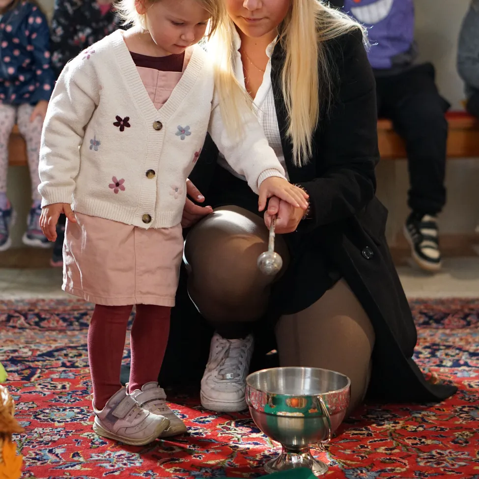 Eine Frau und ein kleines Mädchen knien auf einem Teppich, das Mädchen hält einen kleinen silbernen Becher. Die Frau gießt etwas aus einer größeren silbernen Schüssel in den Becher.