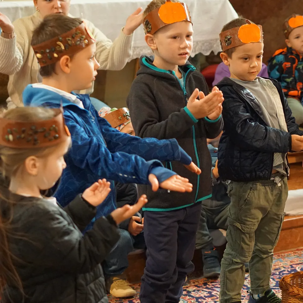 Eine Gruppe von Kindern, die Kopfbänder mit orangefarbenen Kürbissen tragen, klatschen in einem Raum mit den Händen.