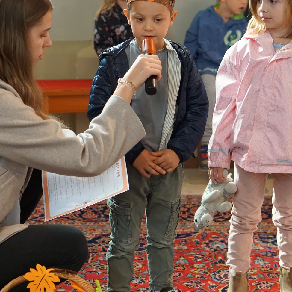 Eine Frau hält ein Mikrofon und ein Papier vor einem jungen Jungen mit einer Kürbis-Mütze, während ein Mädchen ein Kuscheltier hält. Sie stehen auf einem Teppich mit Körben voller Gemüse.