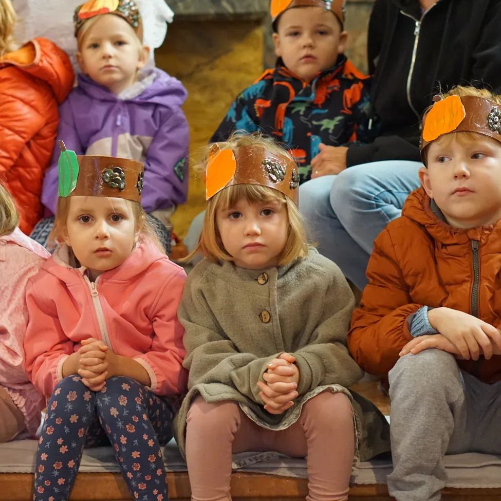 Fünf Kinder sitzen auf einer Bank und tragen orangefarbene Hüte mit Kürbis-Designs. Sie schauen nach vorne, warten wahrscheinlich auf etwas.