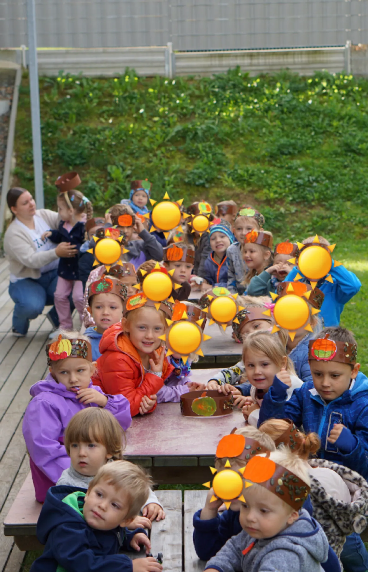 Eine Gruppe von Kindern mit sonnenförmigen Kopfbändern sitzt draußen auf Bänken. Einige halten gelbe Luftballons, und eine Frau steht in der Nähe.