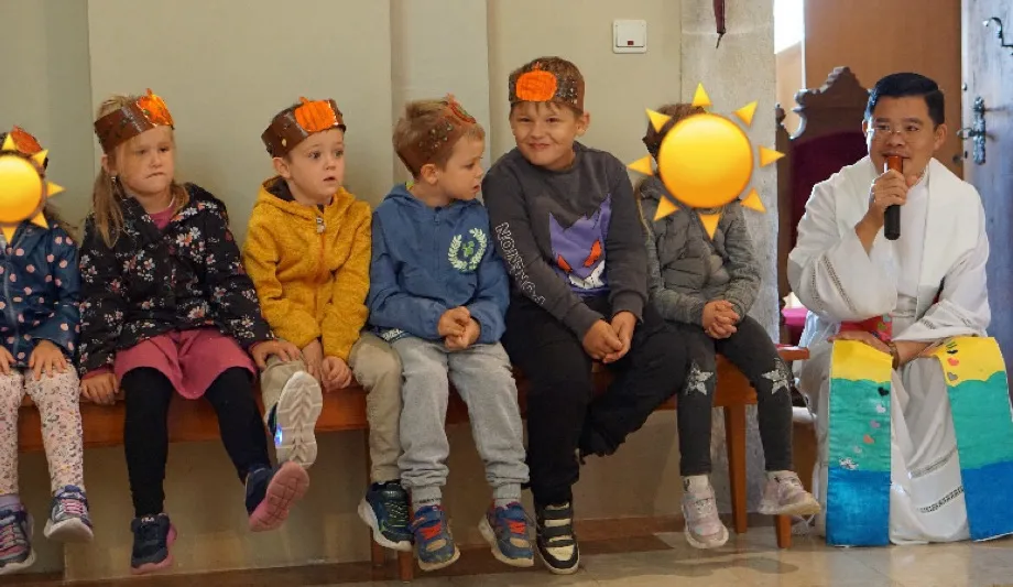 Eine Gruppe von Kindern, die Kronen tragen, sitzen auf einer Bank in einer Kirche. Ein Priester steht vor ihnen und hält ein Mikrofon.