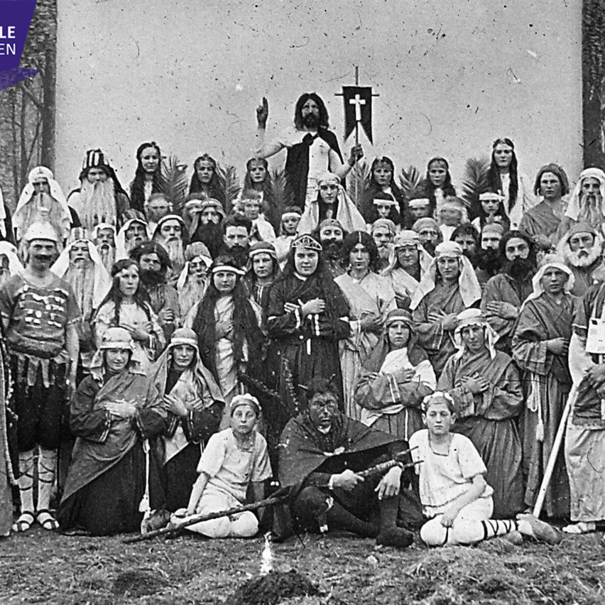 Eine Gruppe von Menschen in altertümlicher Kleidung posiert für ein Foto. Eine zentrale Figur erhebt eine Hand, andere stehen oder sitzen um ihn herum. Die Szene spielt sich im Freien ab.