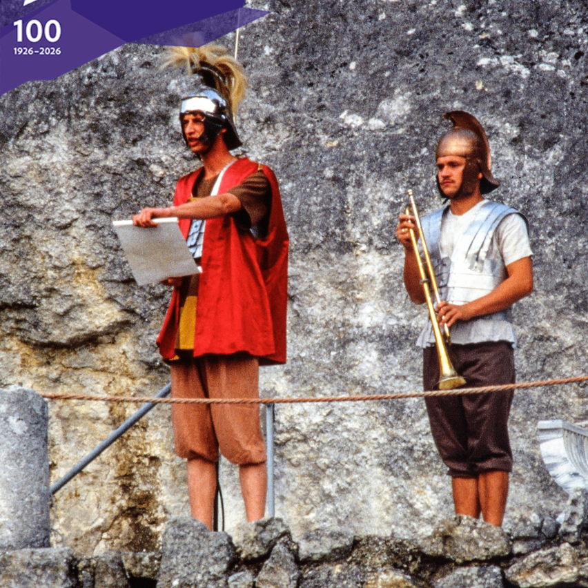 Two men in ancient Roman soldier costumes stand in front of a rock wall. One holds a piece of paper, and the other holds a trumpet. The image has a watermark of '1926-2026'.