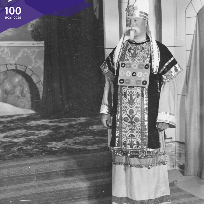 A monochromatic photo shows a man dressed in elaborate ceremonial attire, standing in a grand setting. He wears a tall hat and has a prominent mustache. The background includes an archway and a textured floor.