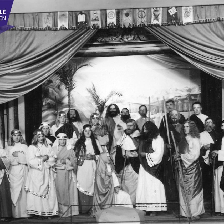 A group of people dressed in white ancient costumes stand on a stage. Some hold staffs. The stage is draped in fabric and adorned with banners. The background shows a scenic view.