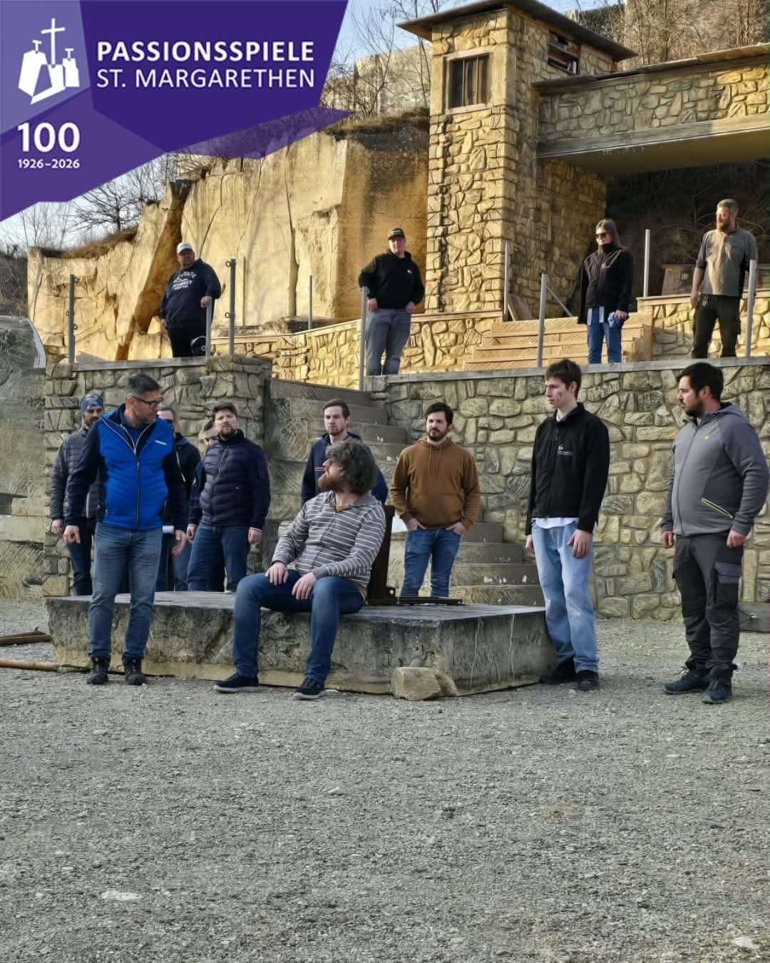 A group of men gather on a stone platform and stairs, some seated, others standing. They wear various jackets and pants, and one man wears a striped shirt. A building with stone walls is behind them.
