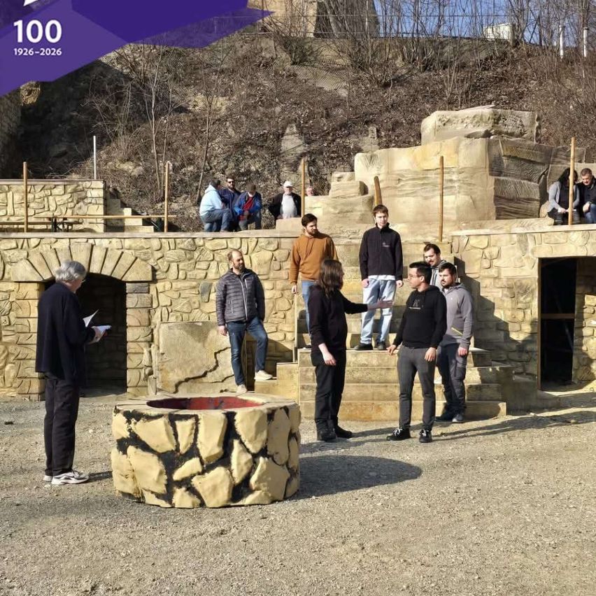 Group of people standing and sitting on stone stairs. One man holds paper. A large stone circle is in the foreground.
