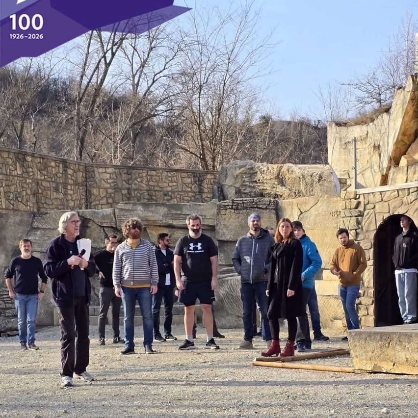 A group of people are gathered in front of a stone structure. A man holds a microphone, likely giving a speech. The scene is set in a historical location, with a number '100' and years '1926-2026' displayed.