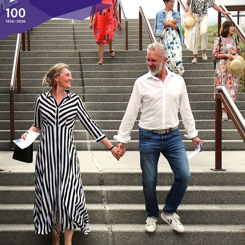 Ein Paar in passenden Outfits hält sich an den Händen, während es eine Treppe hinaufgeht, mit anderen Leuten hinter ihnen. Der Mann trägt ein weißes Hemd und Jeans, während die Frau ein schwarz-weiß gestreiftes Kleid trägt.