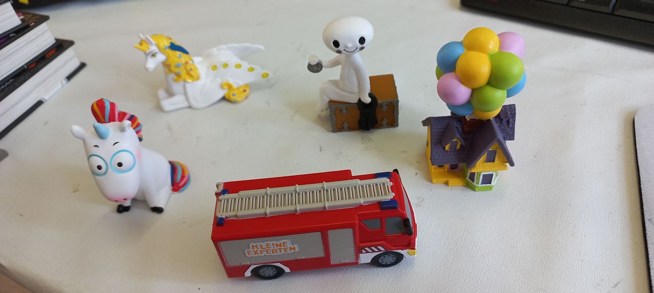 Ein roter Spielzeug-Löschwagen mit der Aufschrift 'Kleine Experten' steht auf einer weißen Oberfläche. In der Nähe sind eine weiße Spielzeugfigur, ein Kasten, ein bunter Luftballon-Strauß und ein kleines Haus.