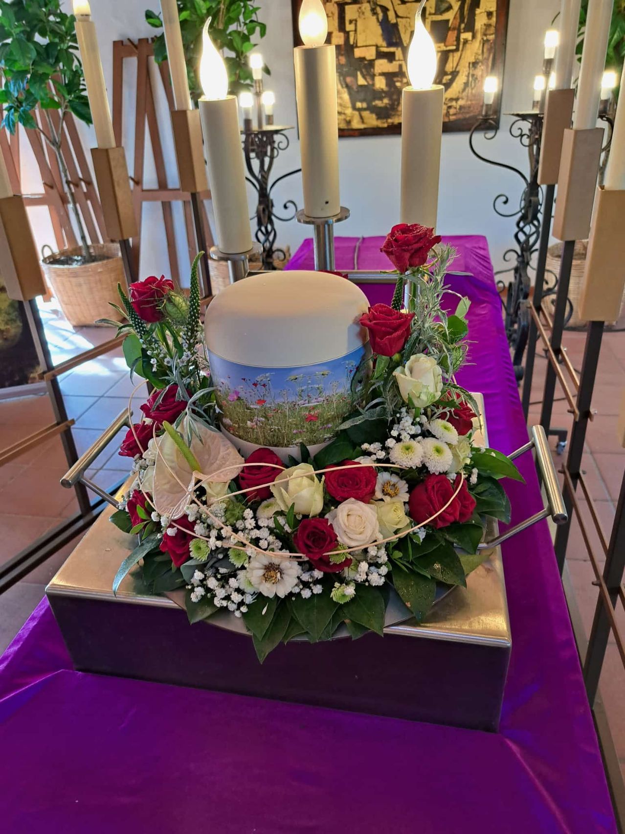 Eine Blumenarrangement mit roten und weißen Blumen umgibt eine zentrale weiße Keramikurne. Die Urne hat ein landschaftliches Design. Das Arrangement steht auf einer silbernen Basis auf einem violetten Tisch.