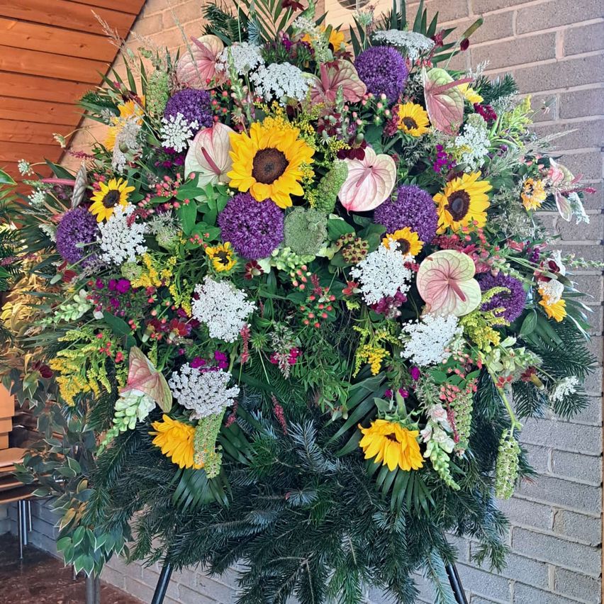 Eine große Blumenarrangement mit Sonnenblumen, lila Blumen und grünen Blättern wird an einer Ziegelwand ausgestellt.