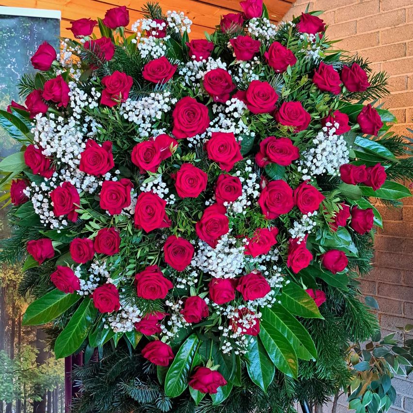 Ein herzförmiges Blumenarrangement mit leuchtend roten Rosen und weißen Babysbreath sowie üppigen grünen Blättern. Das Arrangement wird an einer Ziegelwand präsentiert.