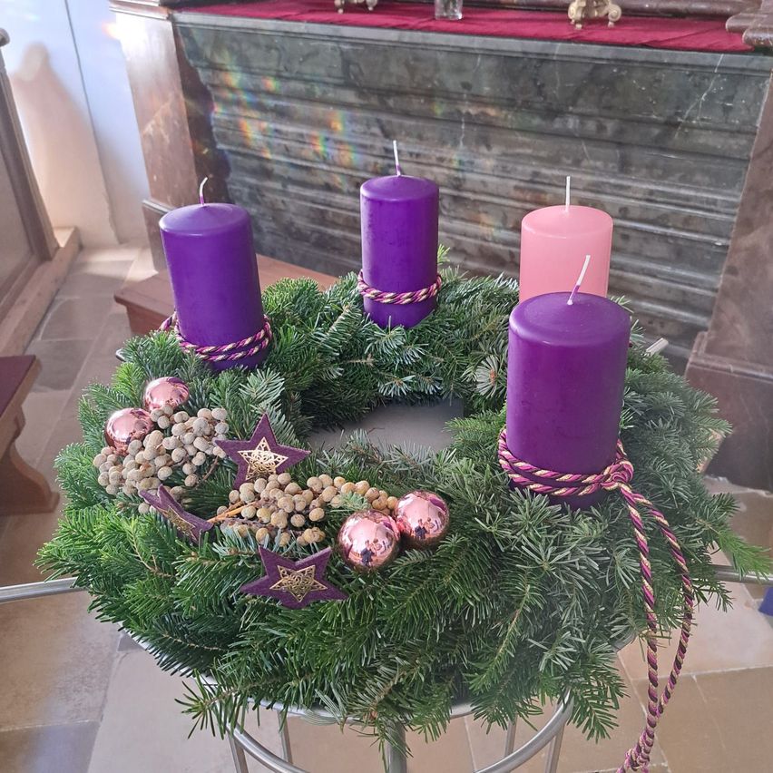 Ein Kranz mit vier lila Kerzen und verschiedenen Ornamenten, darunter Sterne und Kugeln, auf einem Tisch in einer Kirche.