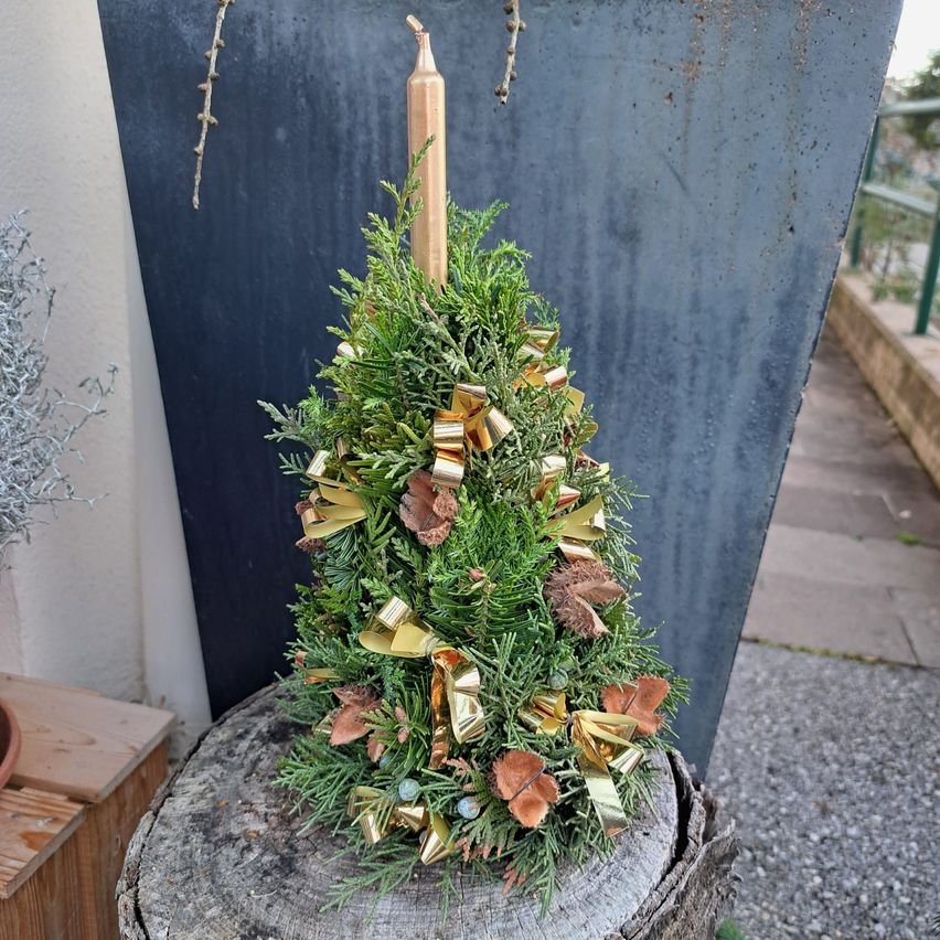 Ein kleiner Weihnachtsbaum mit goldenen Dekorationen und einer hohen Kerze steht auf einem Baumstumpf vor blauem Hintergrund.