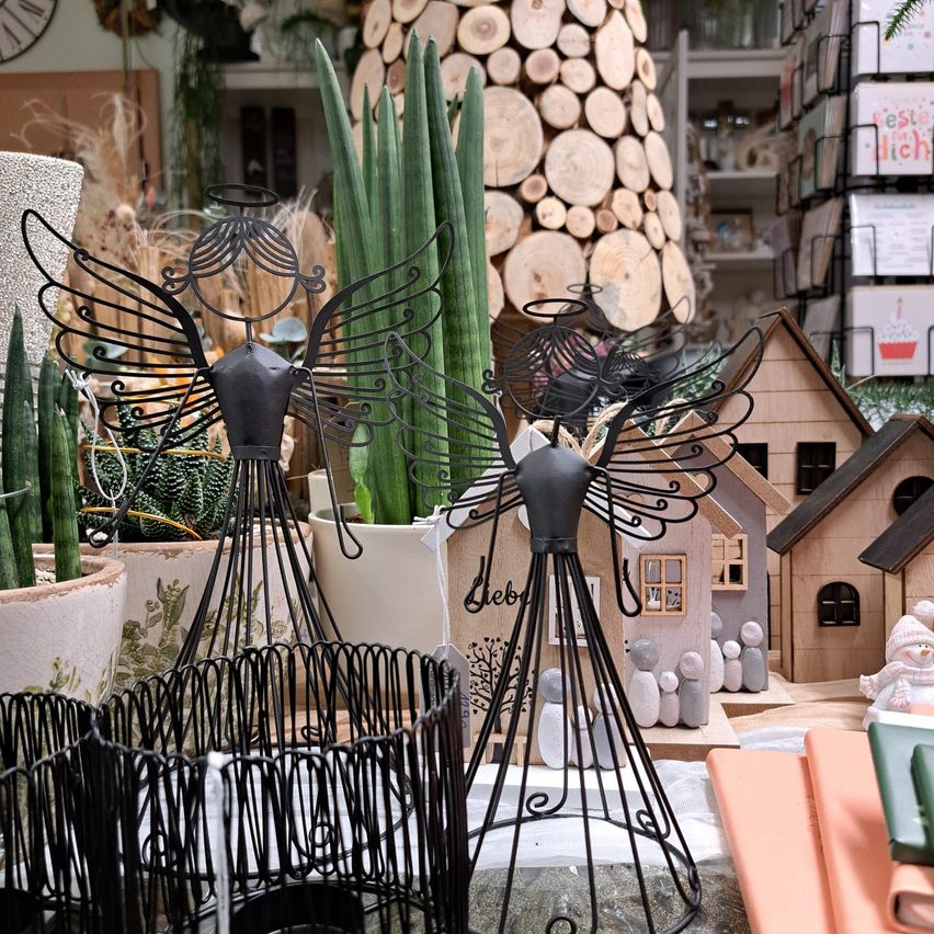 Ein Nahaufnahme von verschiedenen Dekorationsartikeln in einem Geschäft. Zwei schwarze Metallengelfiguren stechen besonders unter Topfpflanzen und Miniaturhäusern hervor.