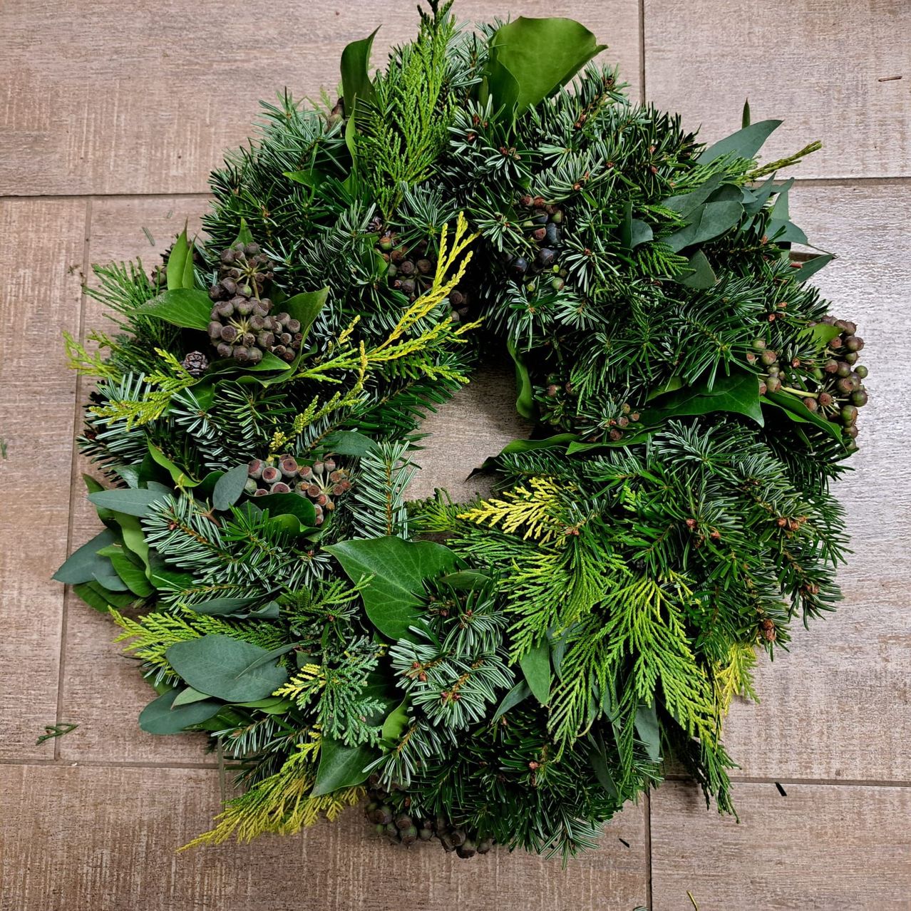 Ein runder Kranz aus immergrünen Zweigen, Nadeln und Beeren auf einer Holzoberfläche.
