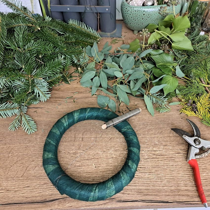 Ein Holztisch zeigt einen gefertigten Kranz mit grünem Laub, einer Metallnadel und Gartenschere. Verschiedene immergrüne Zweige und Blätter sind umher verstreut.