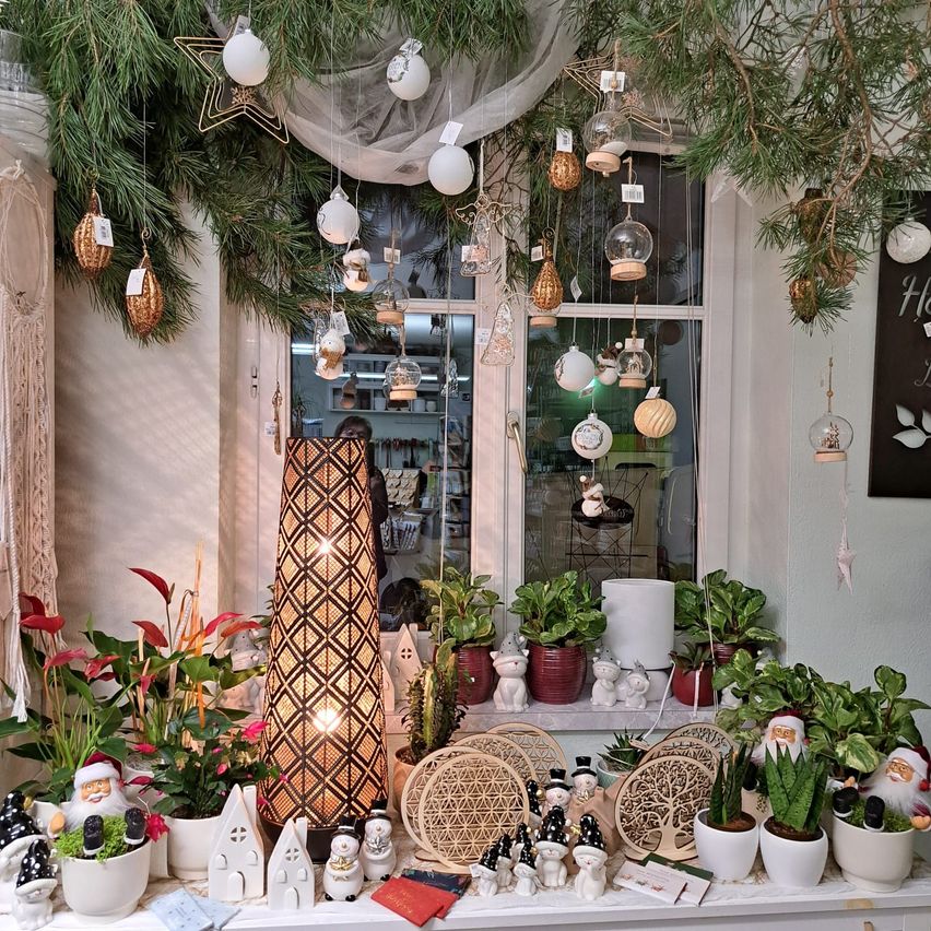 Ein Schaufenster zeigt verschiedene Dekorationen. Weihnachtsbäume mit Ornamenten, eine Lampe und Santa Claus-Figuren zieren die Ausstellung. Verschiedene Pflanzen in Töpfen und Dekorationsartikel sind auf einer weißen Oberfläche angeordnet.
