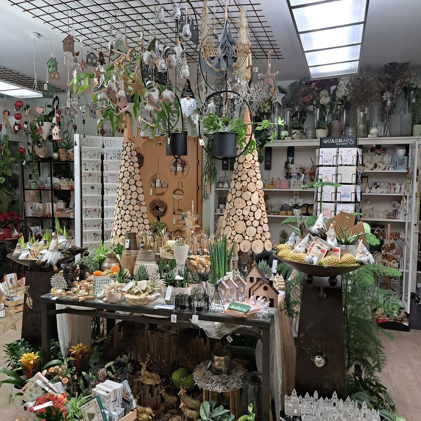 Eine Indoor-Botanik-Ausstellung zeigt einen zentralen Tisch mit verschiedenen Pflanzen, Blumen und Dekorationsartikeln. Umgebende Regale und hängende Dekorationen schmücken den Raum und schaffen eine festliche Atmosphäre.