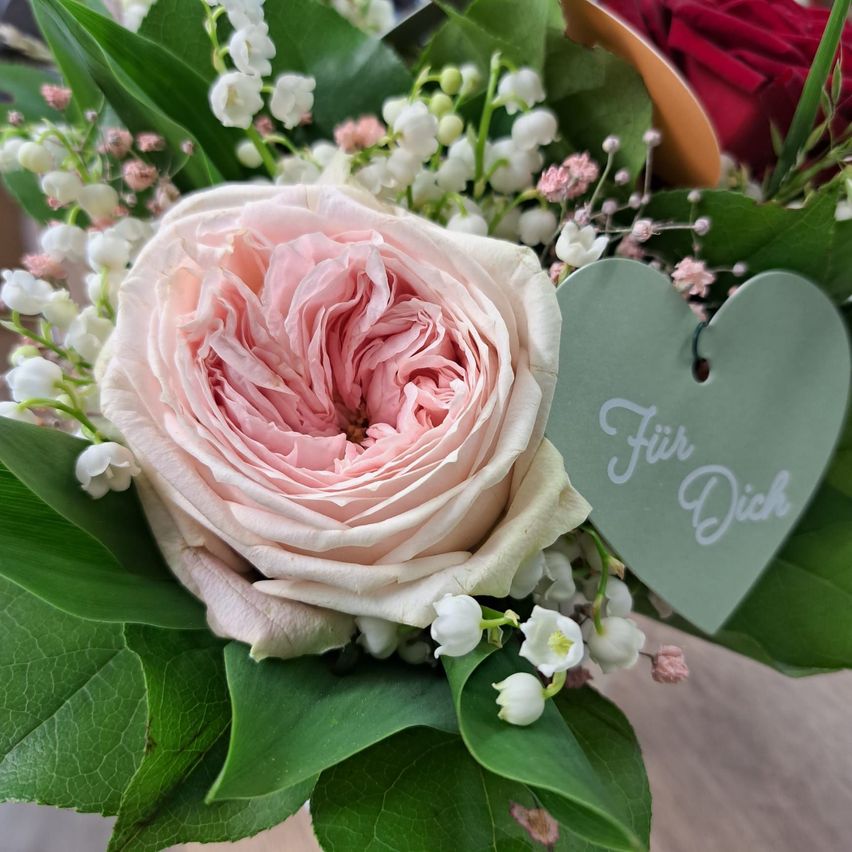 Ein Nahaufnahme einer rosa Rose in einem Strauß mit grünen Blättern und einem herzförmigen Etikett, das 'Für Dich' sagt. Die Rose hat Schichten von Blütenblättern, und der Strauß hat Maiglöckchen und rote Rosen.