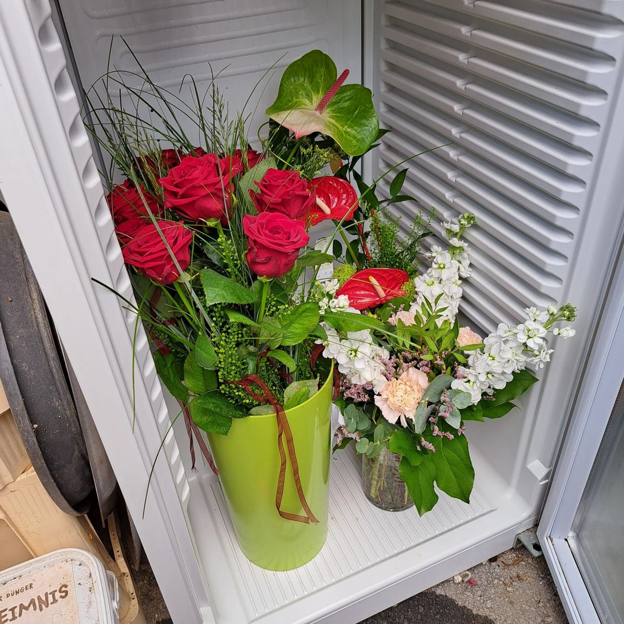 Zwei Vasen mit Blumen stehen in einem offenen Kühlschrank. Die linke Vase enthält rote Rosen und grüne Blätter, während die rechte Vase weiße und rosa Blumen enthält.