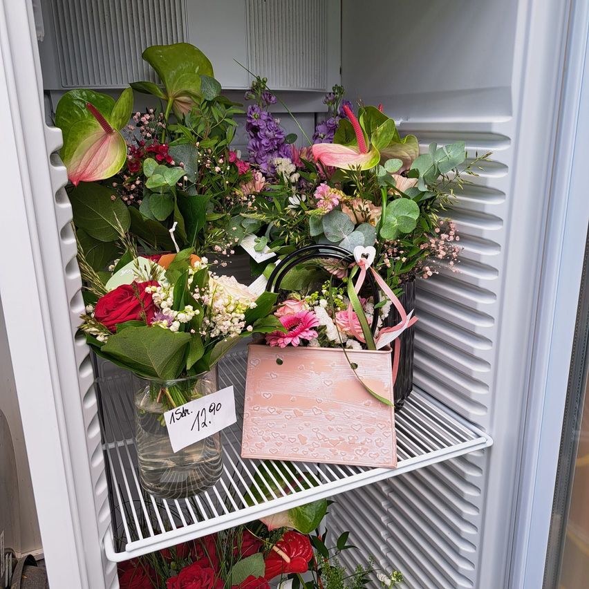Ein Kühlschrank mit Blumen in Vasen und einer rosa Tasche mit Herzdesign, einschließlich eines Straußes mit rosa und lila Blumen, ist auf einem weißen Regal ausgestellt.