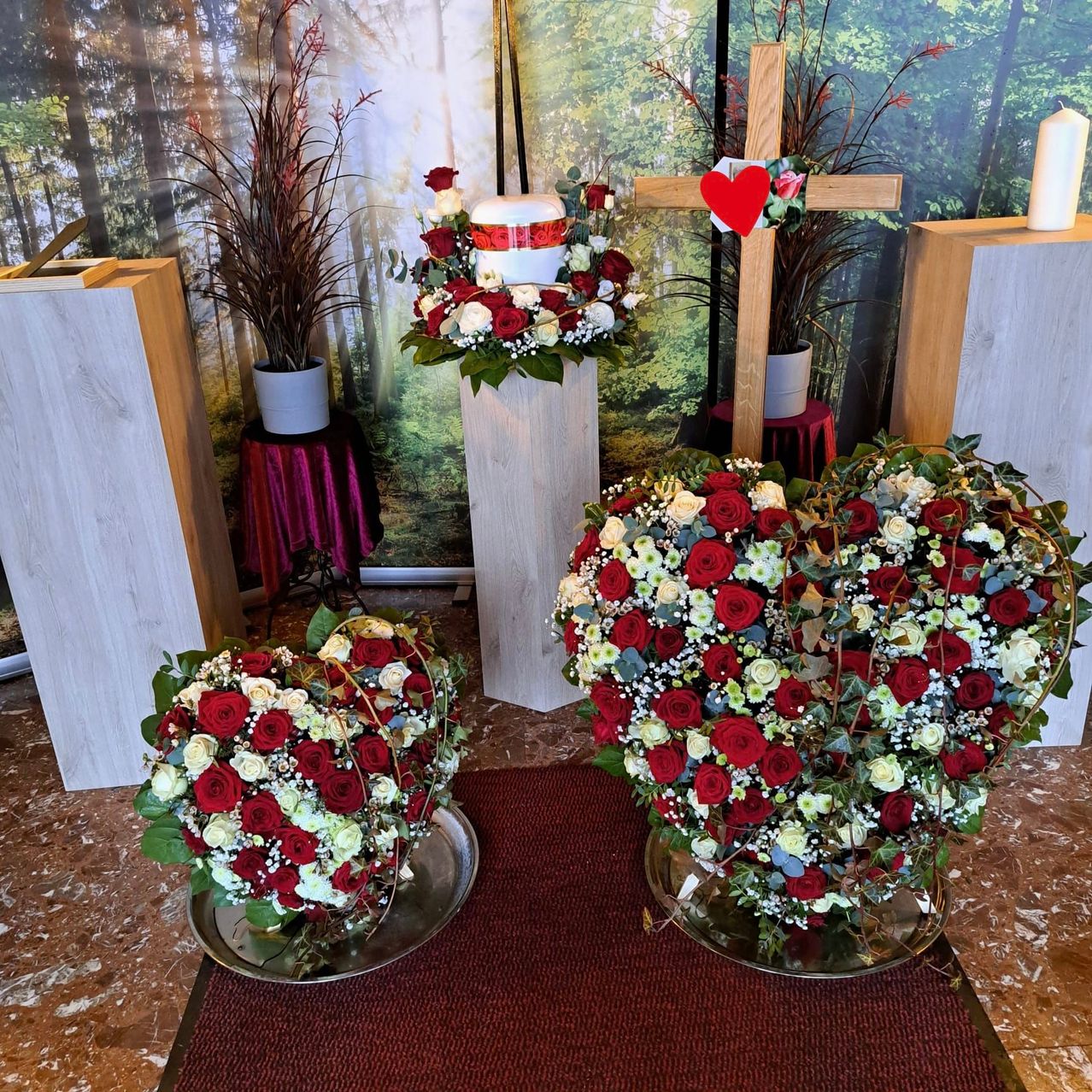 A heart-shaped floral arrangement is displayed on a table, with a wooden cross and a cake nearby. The room is adorned with greenery and has a forest-themed backdrop.