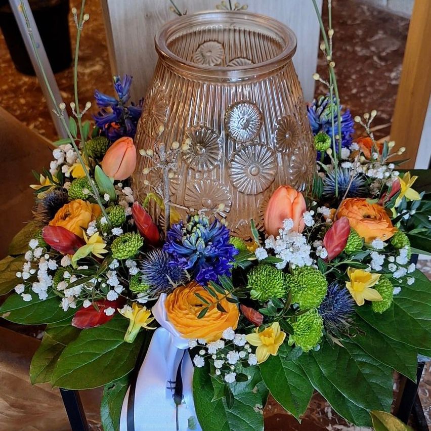 A decorative floral arrangement with various flowers including roses, tulips, and daffodils is displayed in a glass vase. The vase has a floral pattern and is surrounded by green leaves.