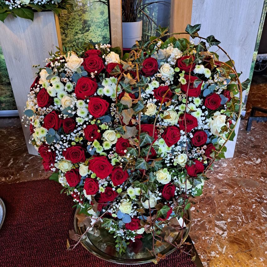 A heart-shaped bouquet of red and white roses with green leaves is on a marble surface, surrounded by other floral arrangements.