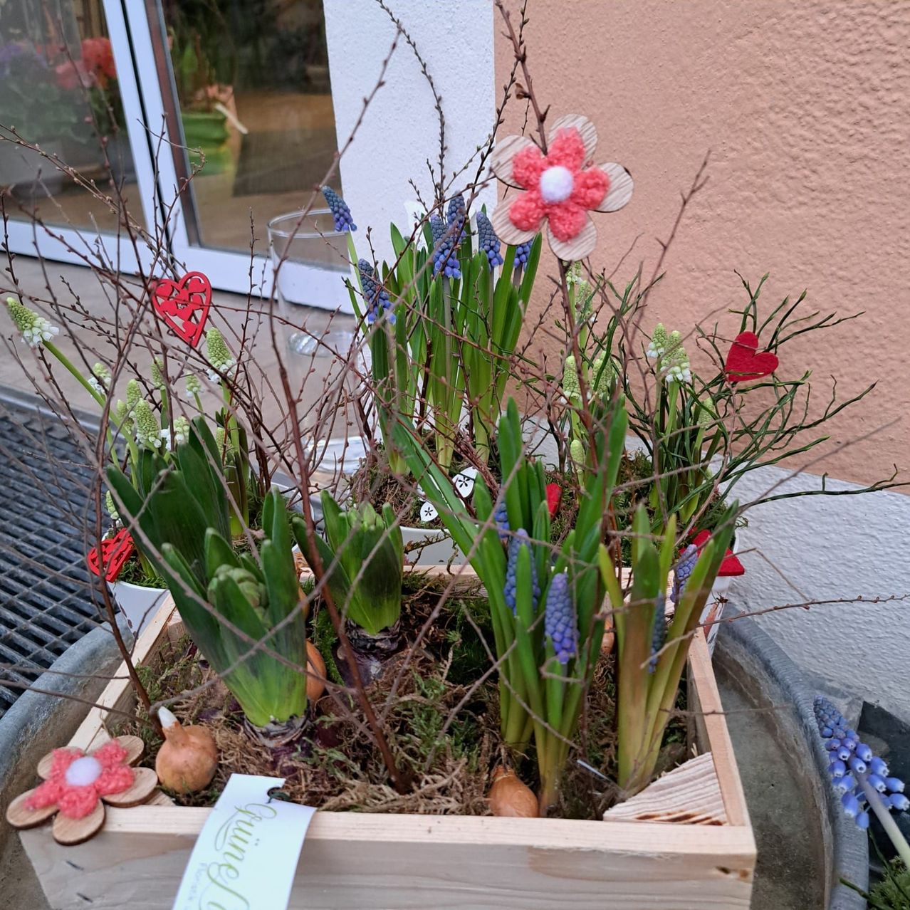 A wooden box contains a spring floral arrangement with green stems, pink crocheted flowers, red hearts, and small onions, placed on a concrete surface.