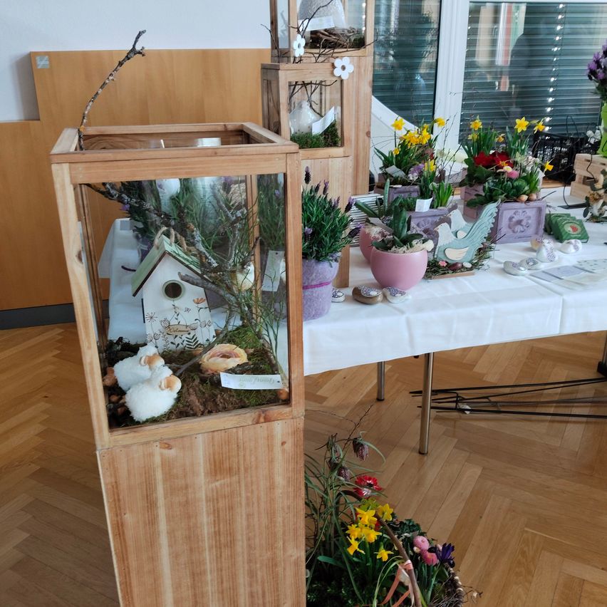 Ein hölzernes Vitrinendisplay mit einem Modellhaus, Schafen und einem Vogelhäuschen im Inneren. Ein Tisch mit weißer Tischdecke ist mit verschiedenen Blumenarrangements und Dekorationsartikeln gefüllt.