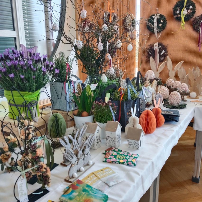 Ein mit einem weißen Tuch bedeckter Tisch, gefüllt mit verschiedenen Dekorationsartikeln wie Ostereiern, kleinen Hasenstatuen, Topfpflanzen und Blumenarrangements. Der Hintergrund zeigt eine strukturierte Wand mit hängenden Kränzen und dekorativen Objekten.