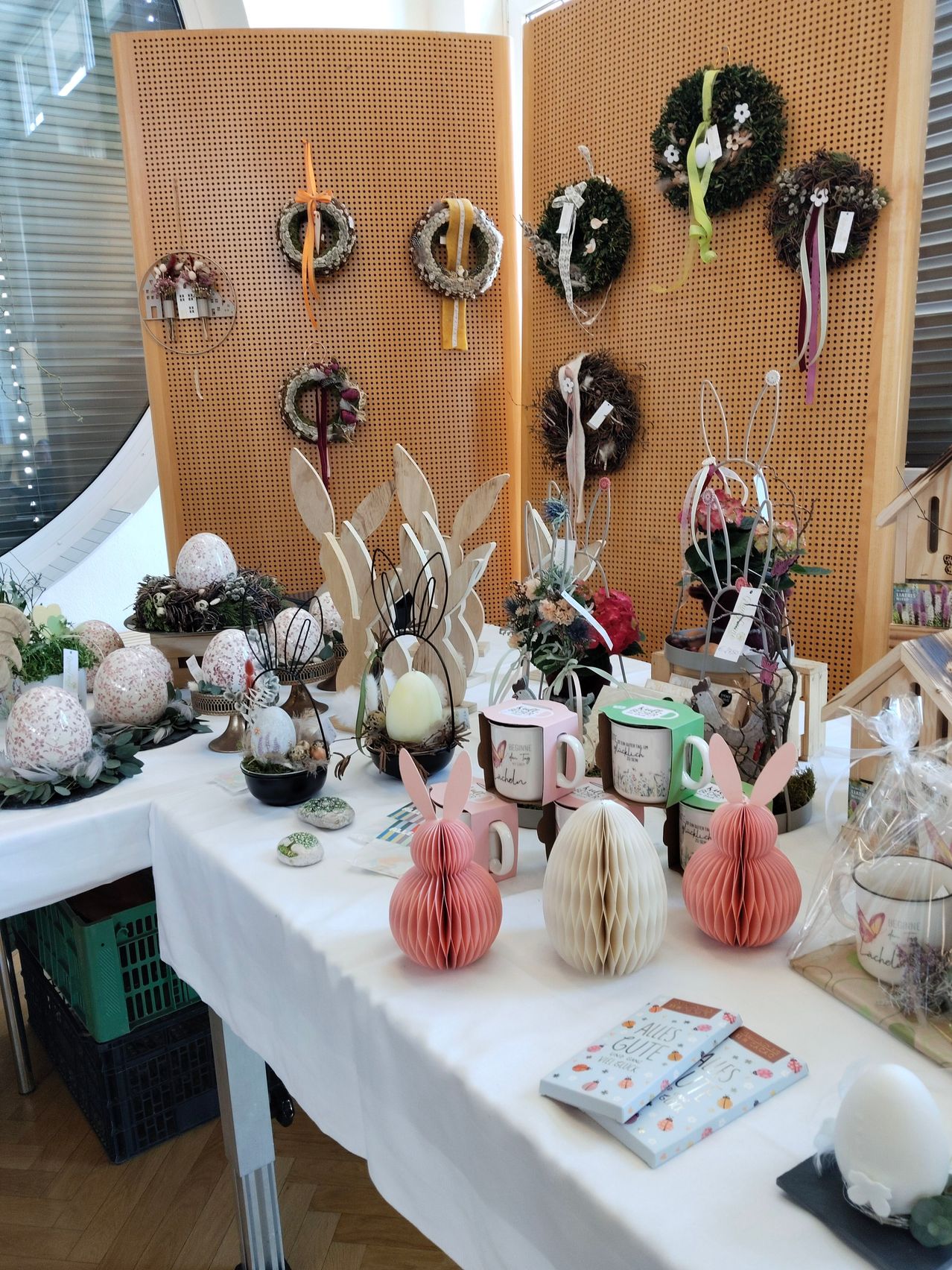 Ein Tisch mit einer weißen Tischdecke ist mit Osterdekorationen gefüllt, darunter dekorative Eier, Hasenfiguren und Blumenarrangements. Hinter dem Tisch ist eine Wand mit Kränzen und Blumenarrangements dekoriert.