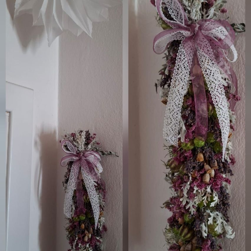 Zwei dekorative Blumenarrangements mit lila Bändern hängen an einer weißen Wand. Das rechts hat einen größeren Bogen und mehr Blumen.