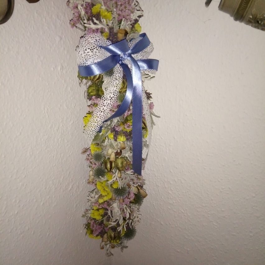 Eine dekorative Blumenarrangement mit getrockneten Blumen und einem blauen Band hängt an einer weißen Wand.