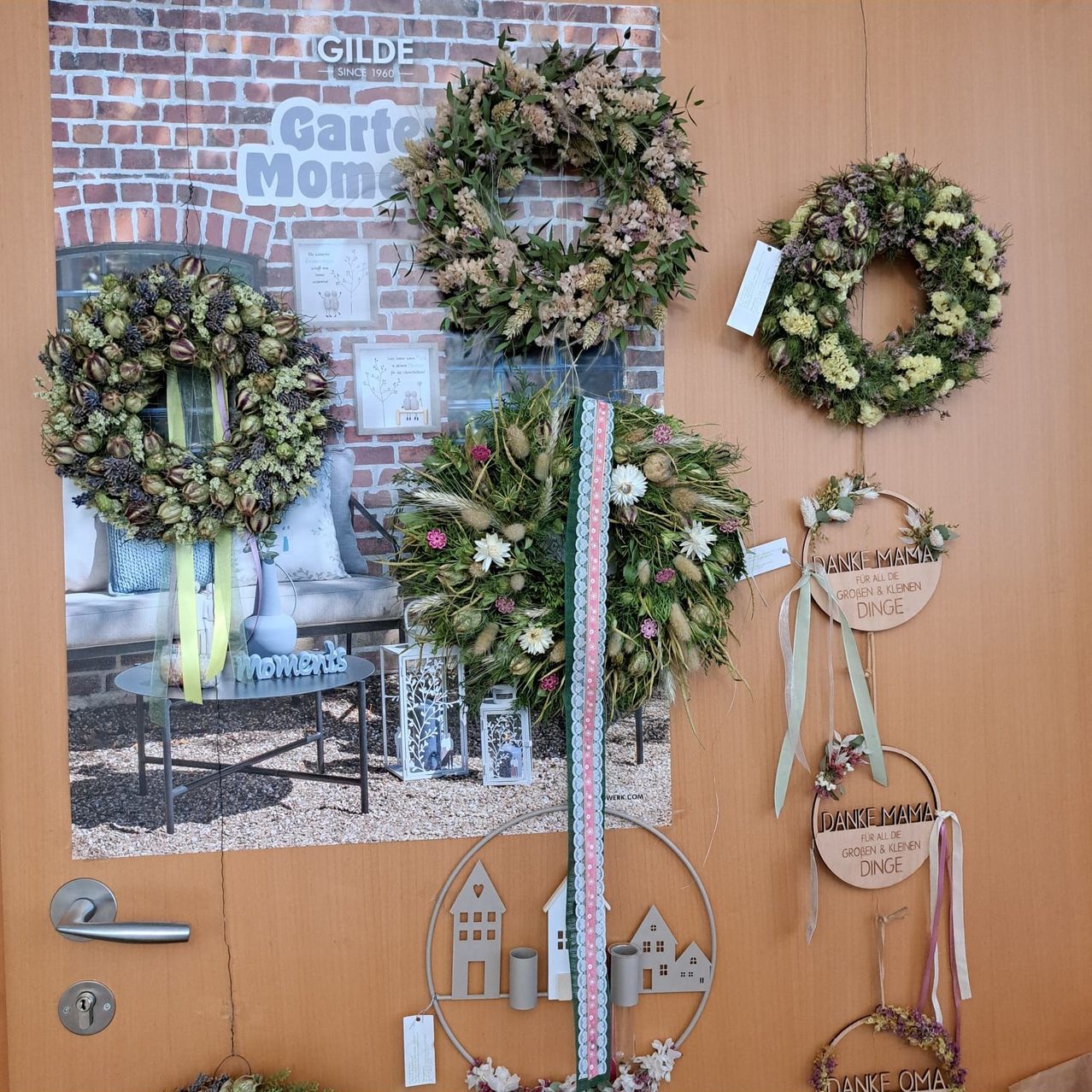 Eine Holztür mit Kränzen und Dekorationsartikeln, die daran hängen. Die Kränze bestehen aus getrockneten Blumen und Blättern. Die Tür hat eine Glasscheibe, die eine Ziegelwand und eine Couch reflektiert.