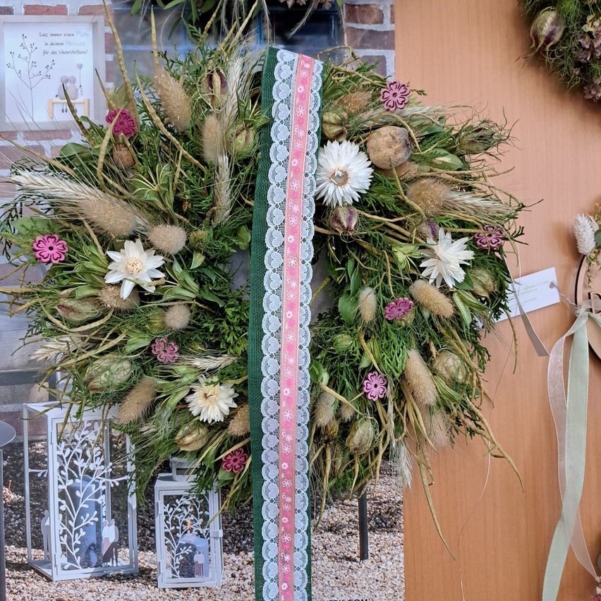 Ein Kranz aus getrockneten Blumen und Grünpflanzen mit einem rosa Band und Spitze, ausgestellt an einer Holzwand.