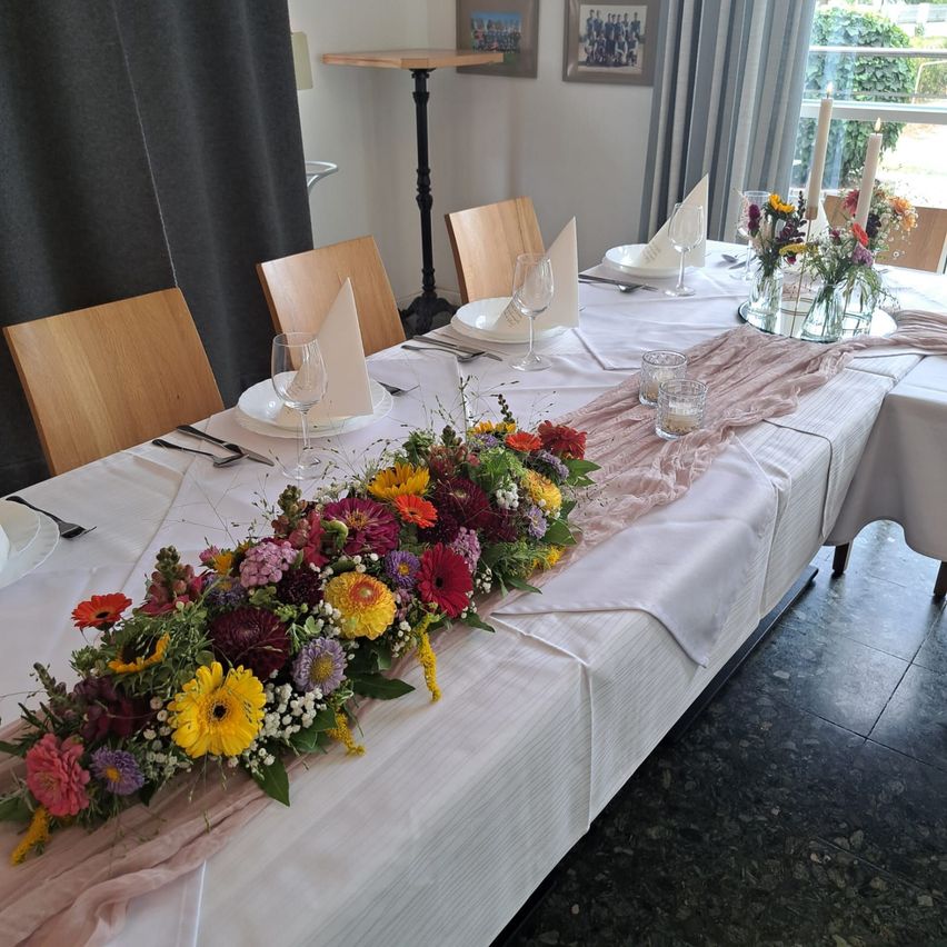 Ein langer Esstisch ist mit Blumen, Tellern, Gläsern und Servietten gedeckt. Der Tisch ist mit einer weißen Tischdecke bedeckt und hat vorne einen rosa Stoff.