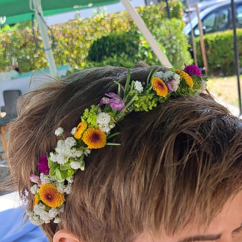 Ein Nahaufnahme von einem Kopf, geschmückt mit einer bunten Blumenkrone, bestehend aus weißen, gelben und lila Blumen. Der Hintergrund ist unscharf mit einem grünen Zelt und Grünpflanzen.